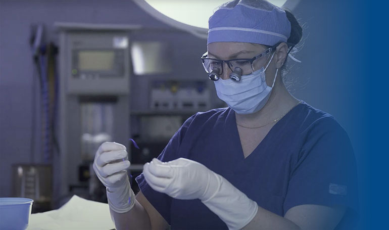 A medical professional wearing scrubs, a mask, and surgical loupes prepares equipment in an operating room.